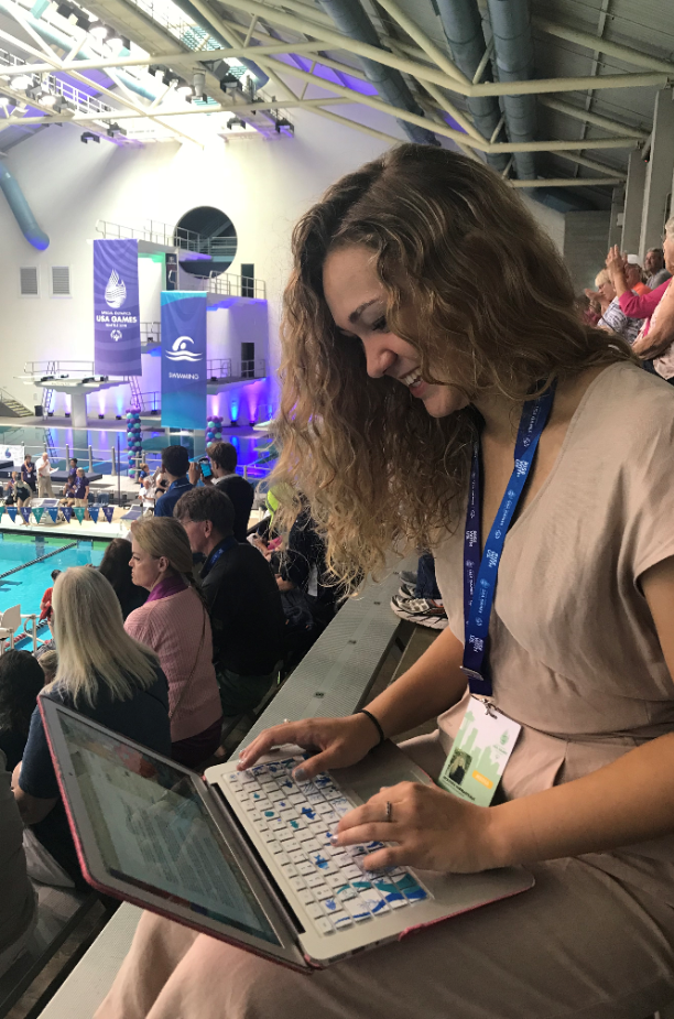 McKenna sits in the stands, typing on her laptop, as swimmers prepare to race.